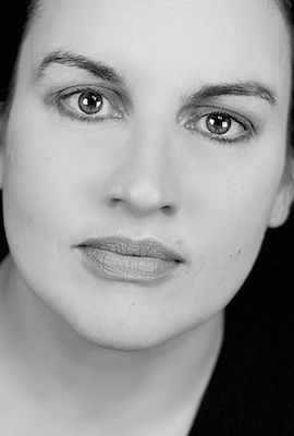 Das Portrait zeigt Verena Mackenberg mit langen, blonden Haaren und einem freundlichen Lächeln. Sie trägt eine weiße Bluse und ihre Hand fasst sich am Hinterkopf ins Haar.